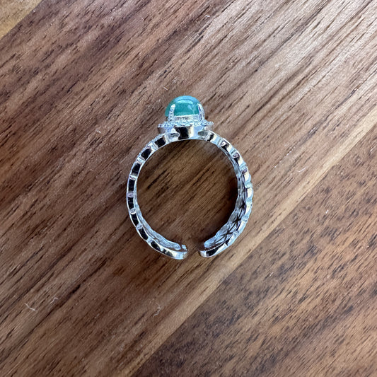 Vintage-inspired openwork band ring with a central green gemstone encircled by a halo of small clear crystals on a wooden background.
