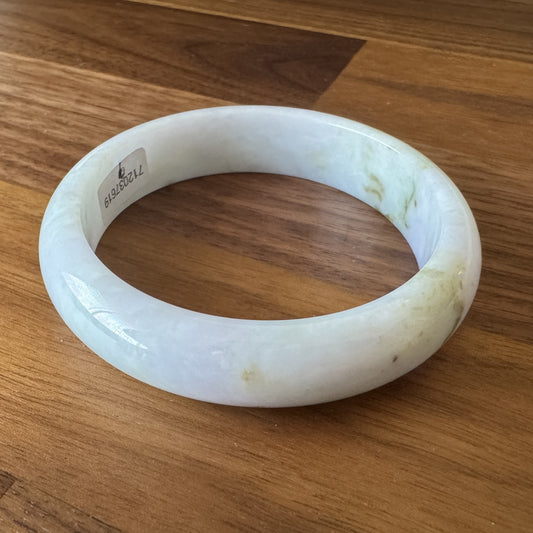 Polished jadeite bangle with translucent white and green hues, showcasing natural marbling patterns, positioned on a wooden surface.