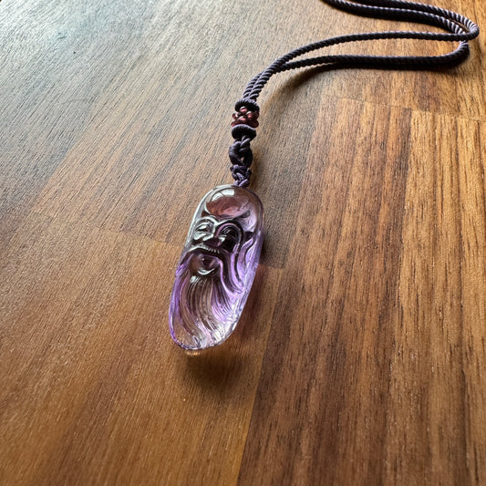 Carved amethyst pendant with sage face design on a braided cord, laid on a wooden surface.