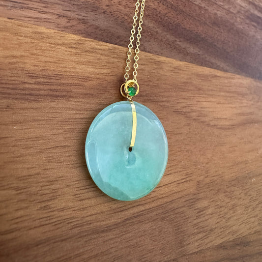 Jadeite disc pendant with gold-tone bail and green gemstone on a golden chain lying on a wooden surface